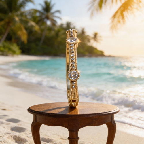 Gold and diamond stackable bracelet displayed upright on a small wooden table at the beach, with turquoise ocean water, white sand, and palm trees softly blurred in the background.