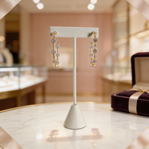 Gold and pavé diamond drop earrings displayed on a white jewelry stand atop a marble surface, set inside a softly lit luxury jewelry boutique with glass cases and a velvet ring box in the background.