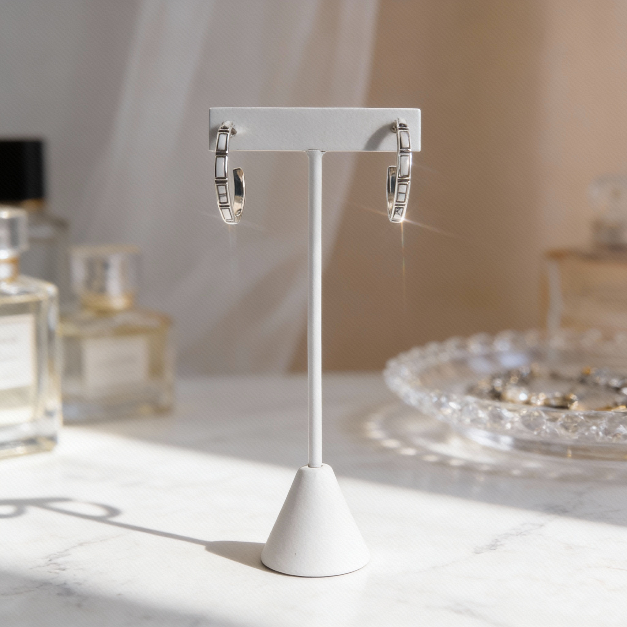 Silver hoop earrings on a white stand with a blurred background of perfume bottles and jewelry.