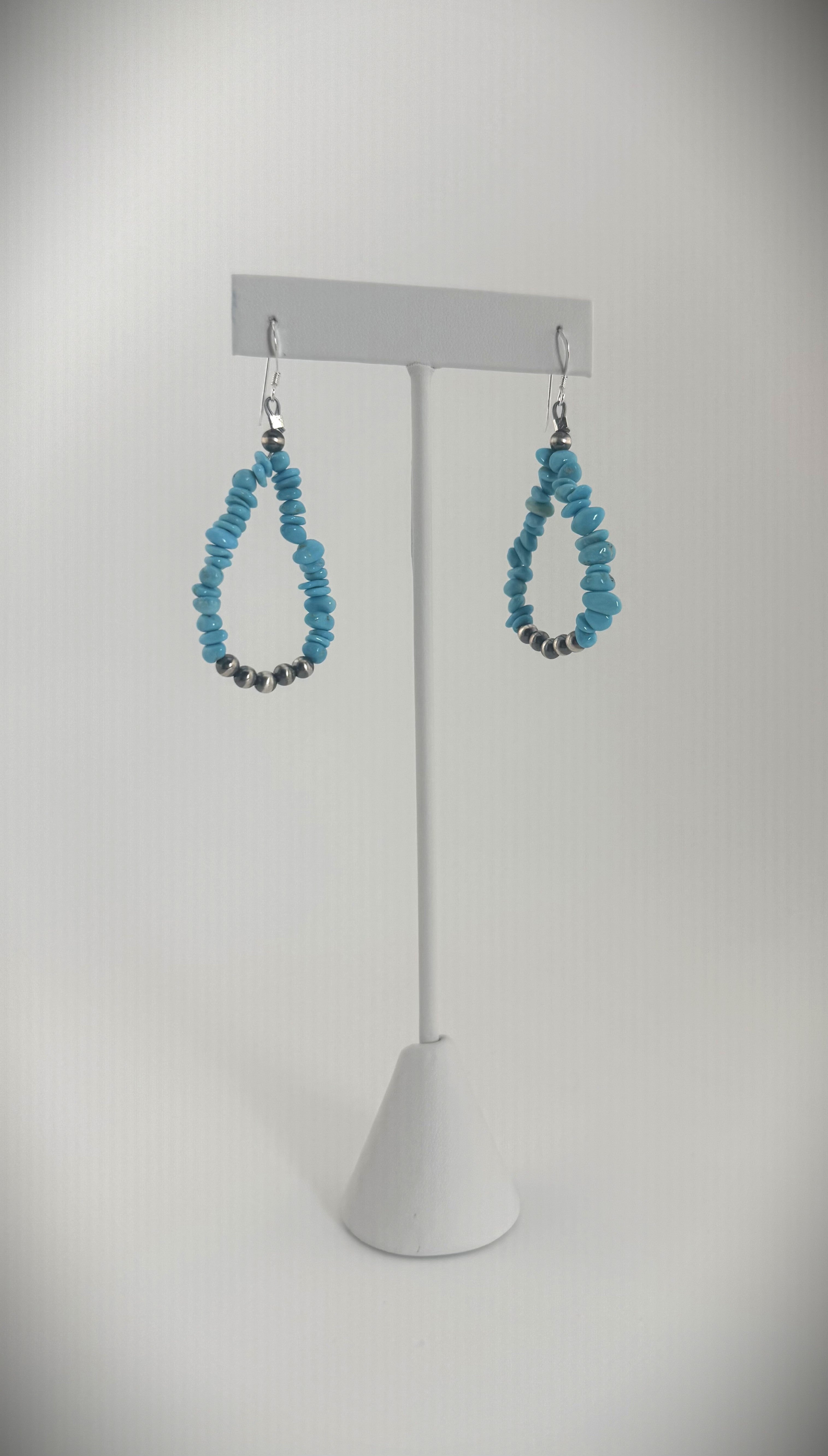 Handcrafted sterling silver teardrop earrings featuring turquoise stone beads, styled on a minimalist jewelry stand with seashell accents